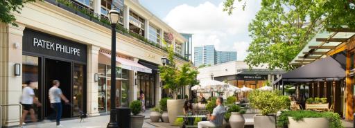 People walking on sidewalks in front of shops, and sitting at tables on a patio, in an outdoor area at Buckhead Village